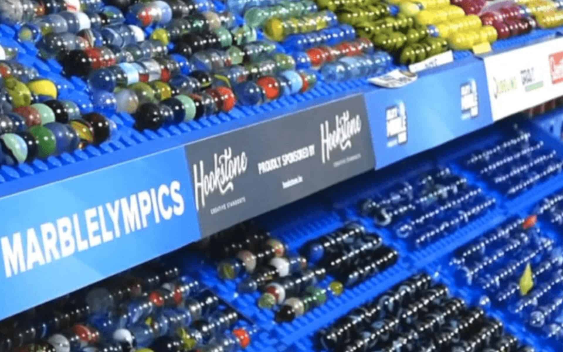 A Marble Display Shows Various Shooters And Alleys In A Blue Plastic Rack With A Sign Reading &Quot;Marblelympics&Quot;